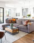 Open-plan living room with gray sectional sofa, colorful cushions, glass coffee table, and kitchen in the background.