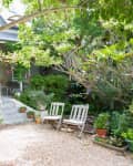 Charming garden with gravel path, three white chairs, lush greenery, and a small porch with potted plants and flowers.