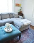 Gray sectional sofa with a white throw, teal ottoman with flowers, and a small plant on a side table in a bright living room.