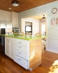 Kitchen with green countertop island, wooden ceiling, white cabinets, and wooden floor, leading to a sunlit room.