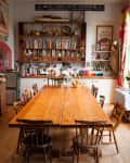 Rustic dining room with a long wooden table, mismatched chairs, bookshelves, and floral centerpiece.