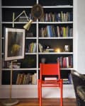 Red chair in front of a black bookshelf filled with books, a gold floor lamp, and a small bust sculpture.