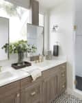 Modern bathroom with dual sinks, gold fixtures, wood cabinets, large mirrors, and a glass shower.
