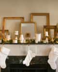 Fireplace mantel decorated with gold frames, candles, and white stockings, surrounded by festive greenery and bottle collection.
