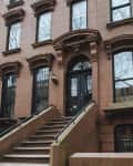 Brownstone building facade with arched doorway, black door, and steps leading up, flanked by wrought iron railings.