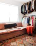 Pink sofa with patterned blankets, red tablecloth, and colorful clothes on racks in a bright room.