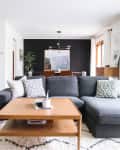 Modern living room with gray sectional sofa, wooden coffee table, and plants, leading to a dining area with dark accent wall.