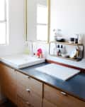 Bathroom vanity with wooden cabinets, large mirror, white sink, and shelf holding toiletries and a clock.