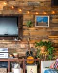 Rustic living room with wood panel wall, mounted TV, string lights, plants, guitar, and vinyl records on a shelf.