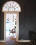 View through arched doorway into dining room with wooden chairs, table, wall clock, and floral arrangement.