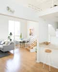 Open-plan living area with a gray sofa, dining table, white staircase, and bar stool on wooden flooring.