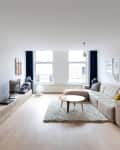 Modern living room with beige sectional sofa, round wooden coffee table, blue dresser, and large windows with dark curtains.