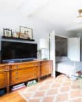 Cozy living room with a wooden dresser, TV, glass coffee table, sofa, and bed, featuring a patterned rug and wall art.