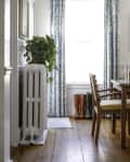 Dining room with wooden floor, radiator with plant, patterned curtains, and three pairs of boots by the window.
