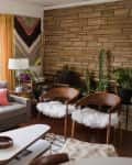 Mid-century modern living room with gray sofa, colorful pillows, wooden chairs, and potted plants against a stone wall.