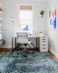 Home office with white desk, swivel chair, abstract art, and blue patterned rug.
