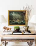 Console table with two lamps, a ship painting, books, a lion sculpture, and a vase with branches.
