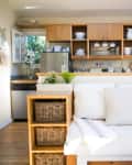 Cozy kitchen with wooden cabinets, open shelving, white dishes, and a sofa with striped pillows and woven baskets.