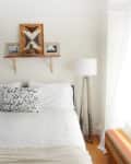 Bedroom with white bedding, abstract pillow, wooden shelf with decor, and a tall floor lamp.