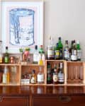 Home bar setup with assorted liquor bottles on wooden shelves, a framed can print above, and a potted plant on the side.