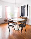 Dining room with round white table, four black chairs, wood floor, red curtains, and abstract wall art.