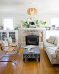 Living room with stone fireplace, wicker chairs, patterned ottoman, and a beige sofa. Bookshelves and plants decorate the space.