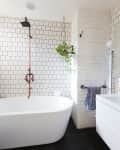 Freestanding bathtub with copper shower, white subway tiles, hanging plant, and towel on a rack in a modern bathroom.