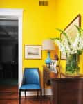 Yellow hallway with a blue chair, wooden sideboard, lamp, framed art, and white flowers in a vase.