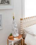Bedroom with white woven headboard, white bedding, side table with statue lamp, and framed art on walls.