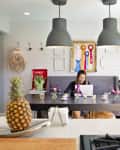 Woman working on a laptop at a dining table with flowers, ribbons on the wall, and a pineapple on the counter.
