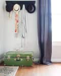 Coat rack with hats and scarves above a green trunk, next to gray curtains in a bright room.