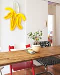 Dining area with wooden table, red and white chairs, potted plant, and yellow banana neon light on the wall.