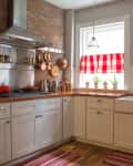 Red buffalo plaid cafe curtains in a white farmhouse kitchen with an apron sink, a stainless steel stove and range hood, an exposed brick wall, and copper pots and pans
