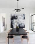 Modern dining room with black table, light wood chairs, abstract art, and glass doors. Black staircase railing on the left.