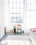 Bright bedroom with large window, green chair, potted plant, white dresser, and cozy bed with pink blanket.