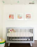 Gray crib with colorful stuffed animals, polka dot bedding, and three abstract paintings on the wall. Green birds on ceiling.