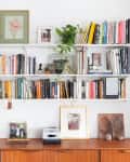 Bookshelf with assorted books, framed art, and plants above a wooden sideboard with photos and a lamp.