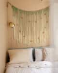 Beige bedroom with macramé wall hanging, cream pillows, and a gold wall lamp.