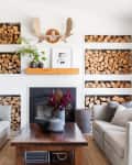 Cozy living room with stacked firewood, white sofas, a wooden coffee table, and a fireplace adorned with moose antlers.
