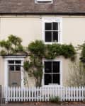 Two-story cream house with white-framed windows, climbing plants, and a white picket fence.