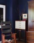 Dark blue room corner with a wooden chair, a cabinet with drawers, and a vase with branches on top.