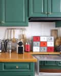 Green kitchen cabinets with coffee machines, cookbooks, and utensils on a wooden countertop.