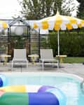 Poolside with colorful inflatable rings, yellow-striped umbrellas, and lounge chairs near a glass greenhouse.