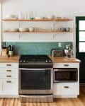 White kitchen with wood countertops, teal backsplash, open shelves, and stainless steel appliances, including a mixer and toaster.
