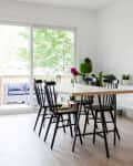Dining room with a wooden table, black chairs, plants, and artwork, next to a sliding glass door leading to a deck with a sofa.