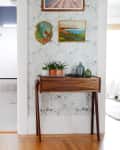 Hallway with wooden console table, potted plants, and framed art on floral wallpaper, leading to a bright room.