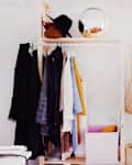 Open wardrobe with hanging clothes, a black hat, a round mirror, and a plant in a bright bedroom.