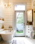 Bathroom with clawfoot tub, chandelier, and glass door leading to garden. White shiplap walls and patterned floor rug.