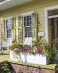 Yellow house porch with green shutters, white planters filled with colorful flowers, and a white bench.