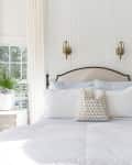 Cozy bedroom with white bedding, patterned pillow, bedside tables, potted plants, and wall-mounted lamps.